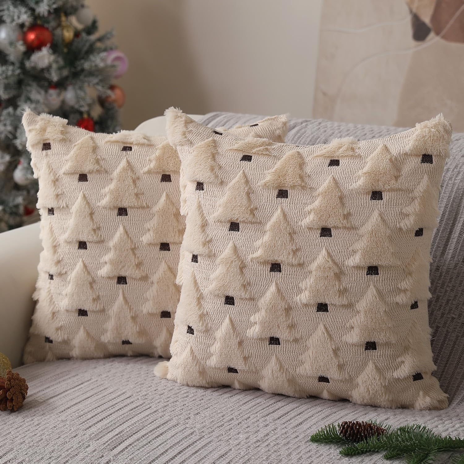 Two beige plush Christmas tree pillow covers with dark brown accents on a sofa, with a decorated Christmas tree in the background.