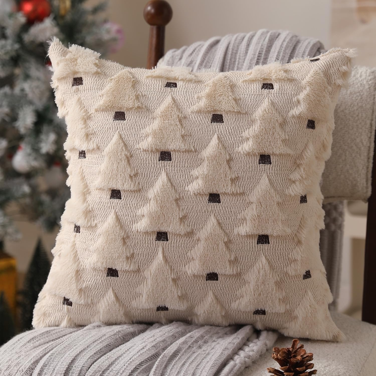 Beige plush throw pillow cover with embroidered Christmas trees, placed on a chair with blankets and a Christmas tree in the background.