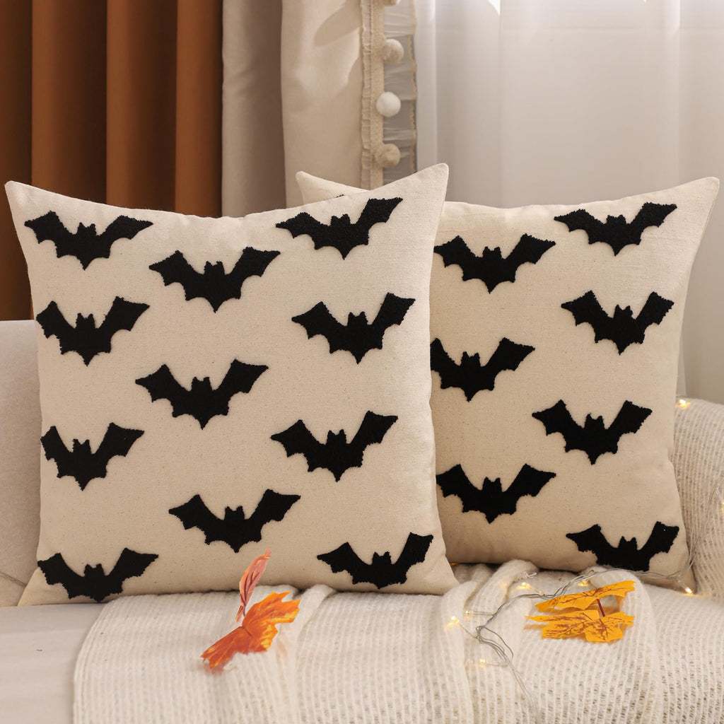 Two cream-colored cotton pillow covers with black embroidered bats, displayed on a sofa with a white knit throw and autumn leaves.