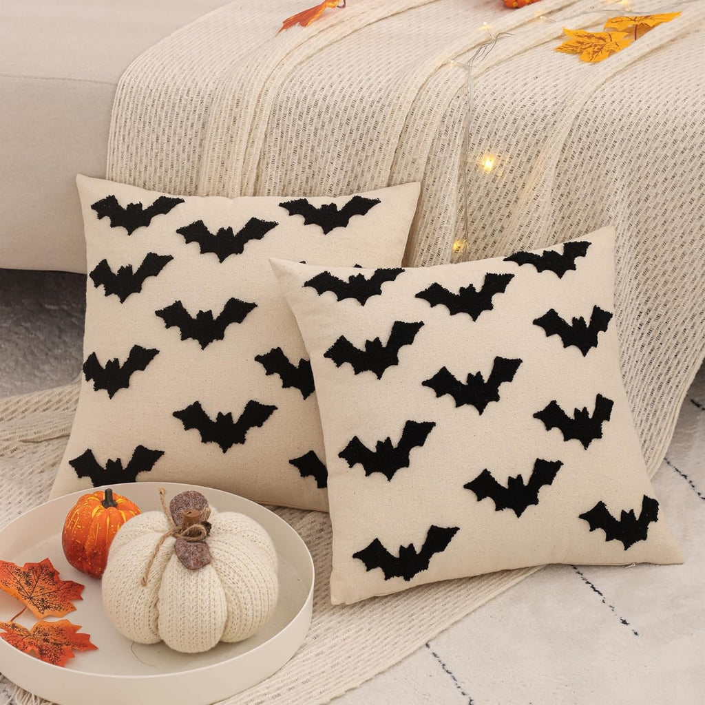 Two cream-colored Halloween pillow covers with black embroidered bats, displayed on a couch with decorative pumpkins and autumn leaves.