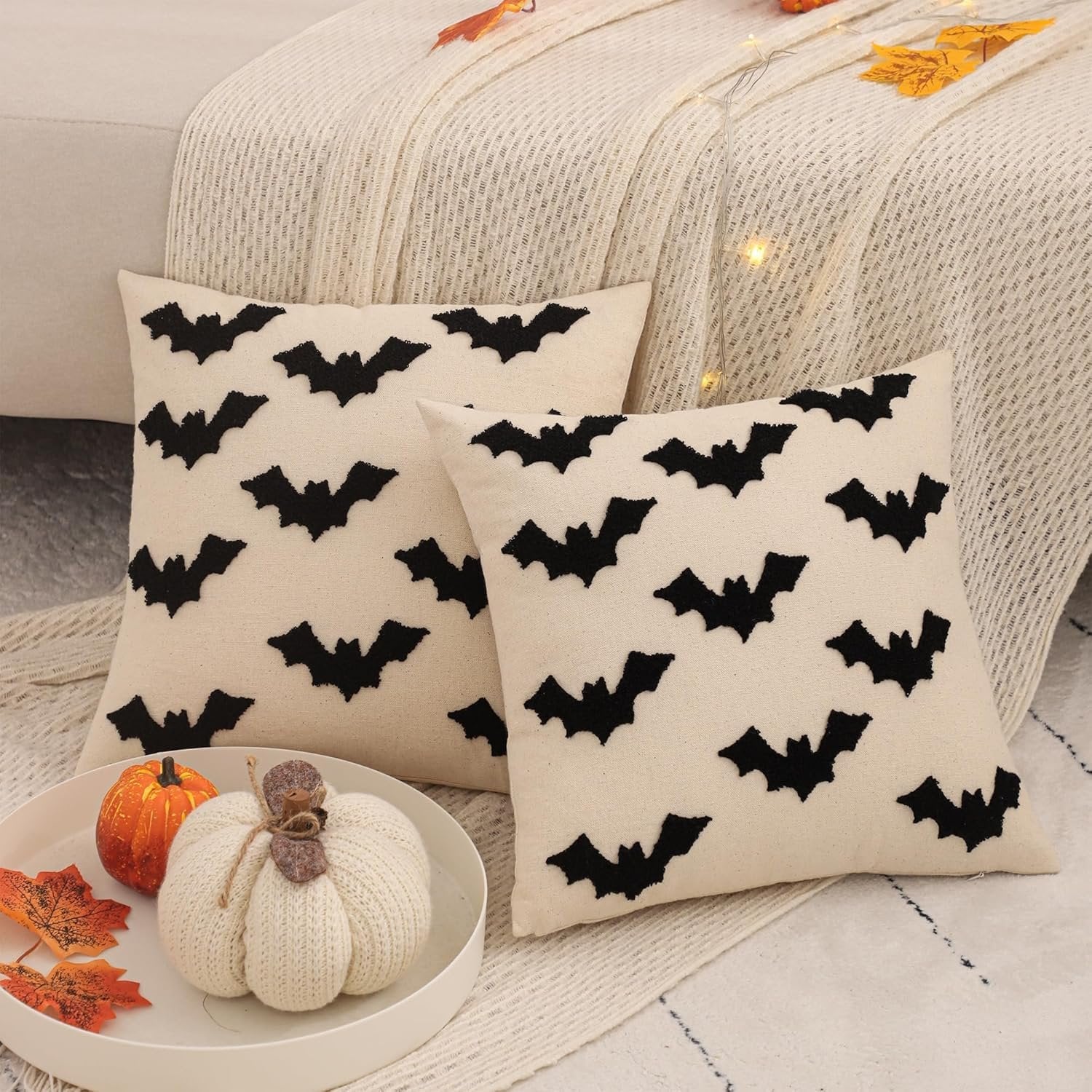 Two cream-colored Halloween pillow covers with black embroidered bats, displayed on a couch with decorative pumpkins and autumn leaves.