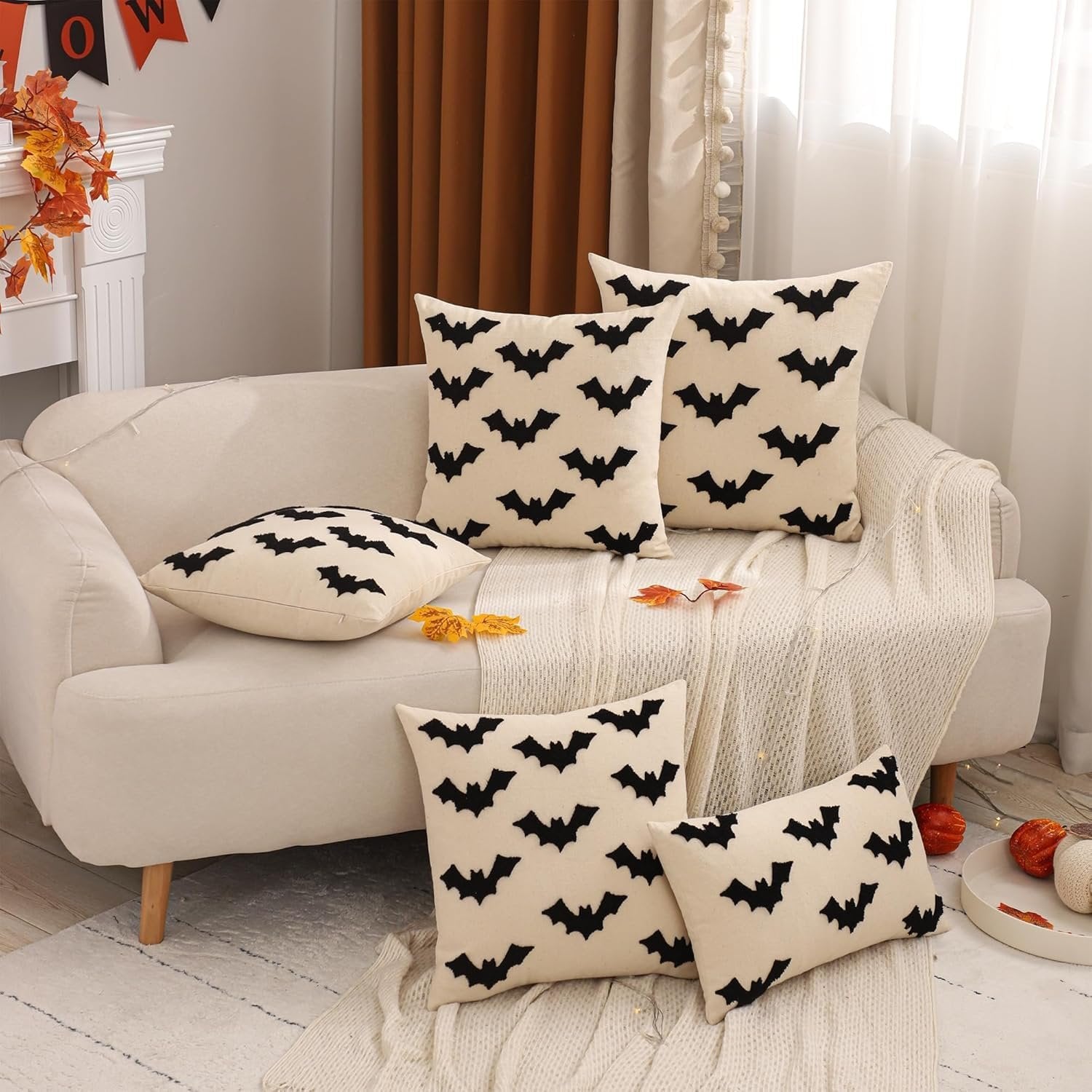 Set of four cream-colored Halloween pillow covers with black embroidered bats, arranged on a sofa with a white throw blanket.