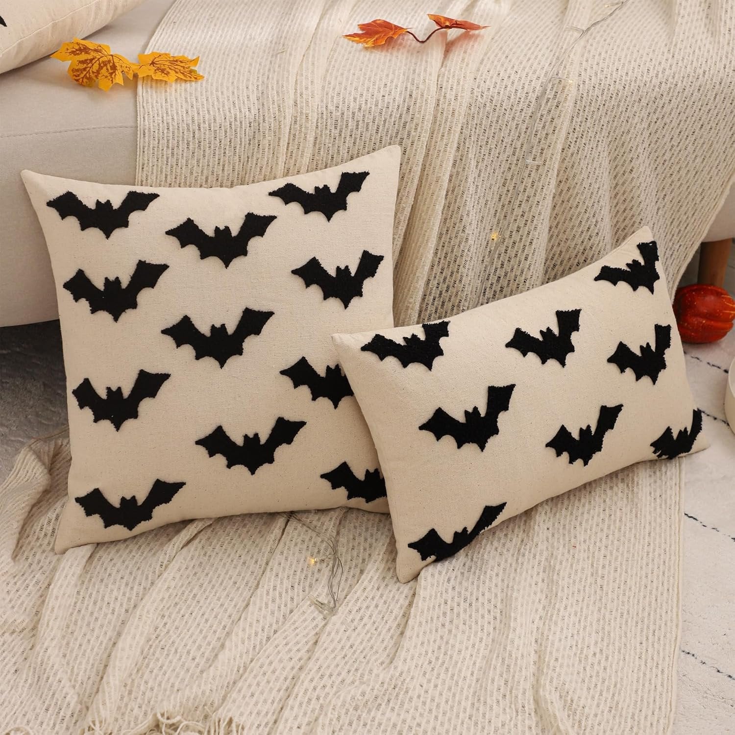 Two cream-colored Halloween pillow covers with black embroidered bats, displayed on a textured throw blanket.