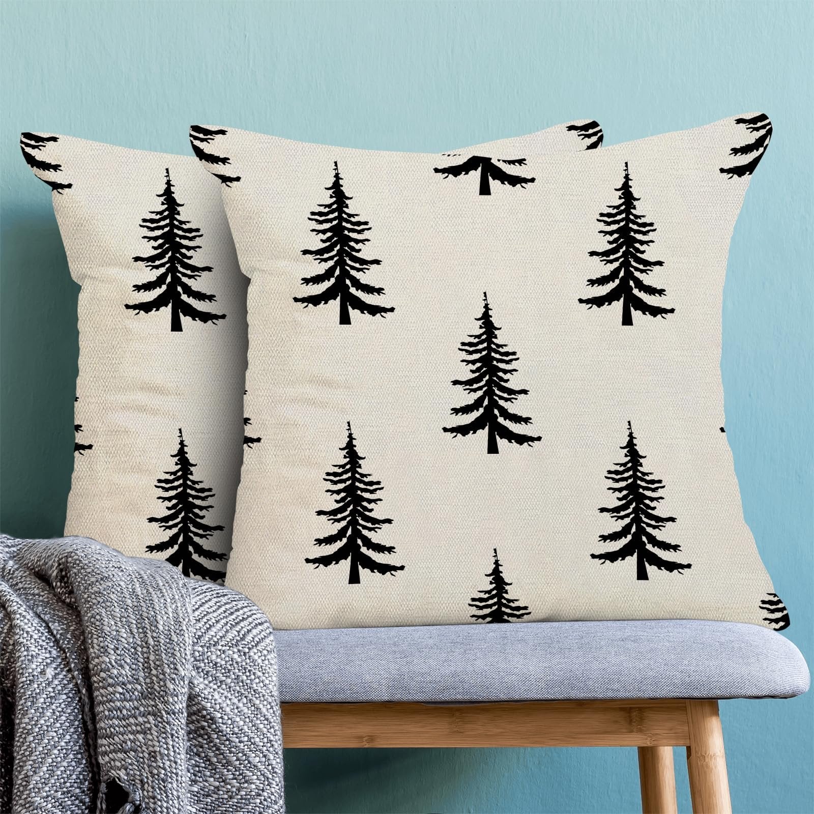Two cream pillow covers with black Christmas tree pattern on a bench with a grey throw blanket.