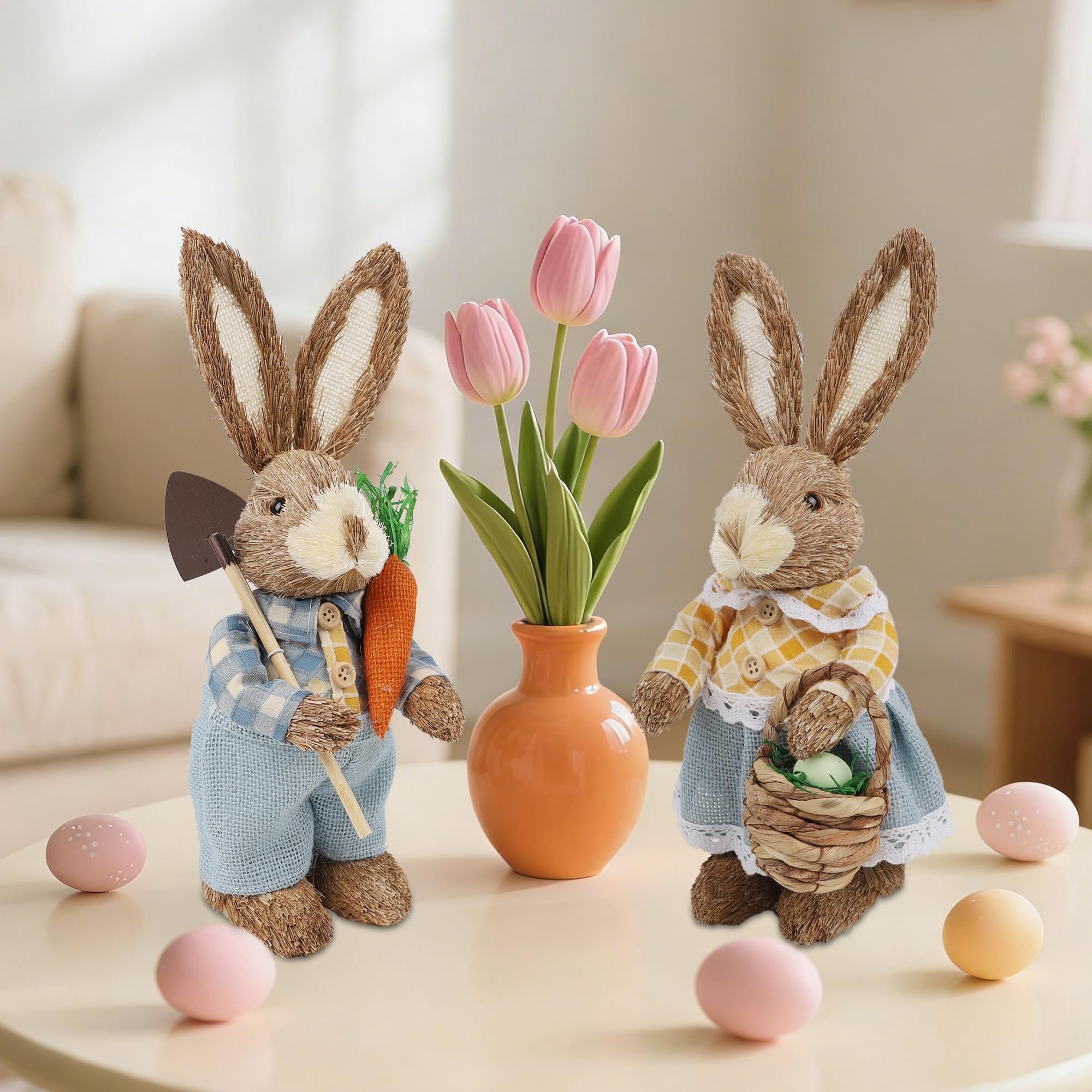 Two straw Easter bunny figurines, one holding a shovel and carrot, the other a basket with eggs, with pink tulips and decorative eggs on a table.