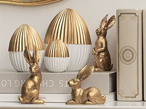 Two gold resin bunny figurines and two gold and white decorative eggs on a shelf with books.