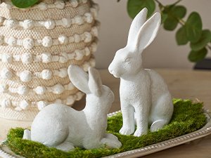 Two white resin bunny figurines on a bed of moss in a decorative tray, with a textured pot in the background.