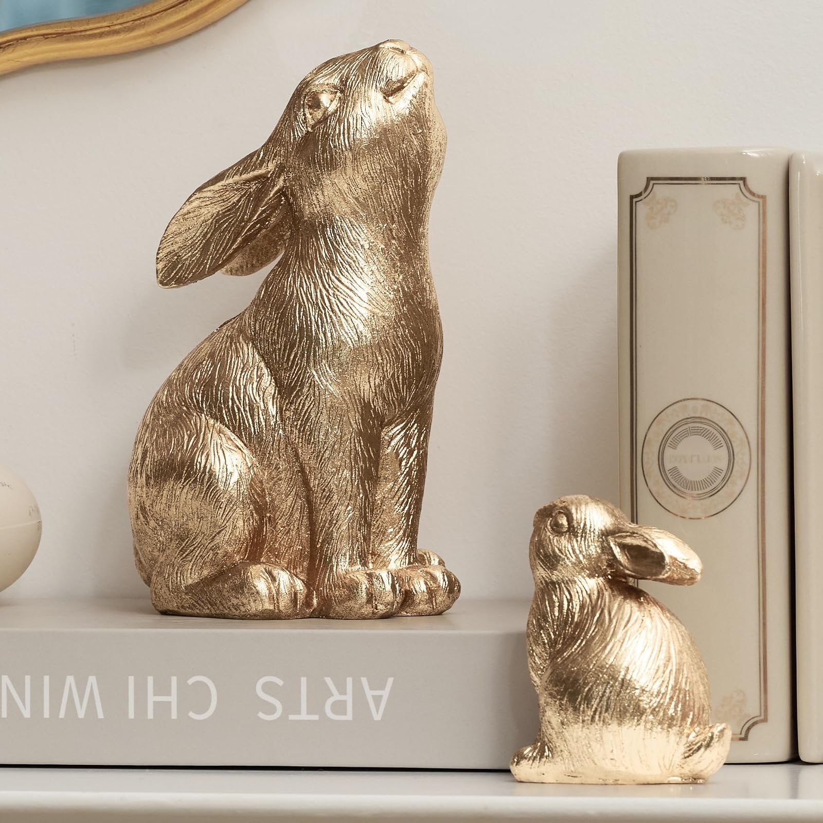 Two gold resin bunny figurines displayed on a shelf with decorative books.