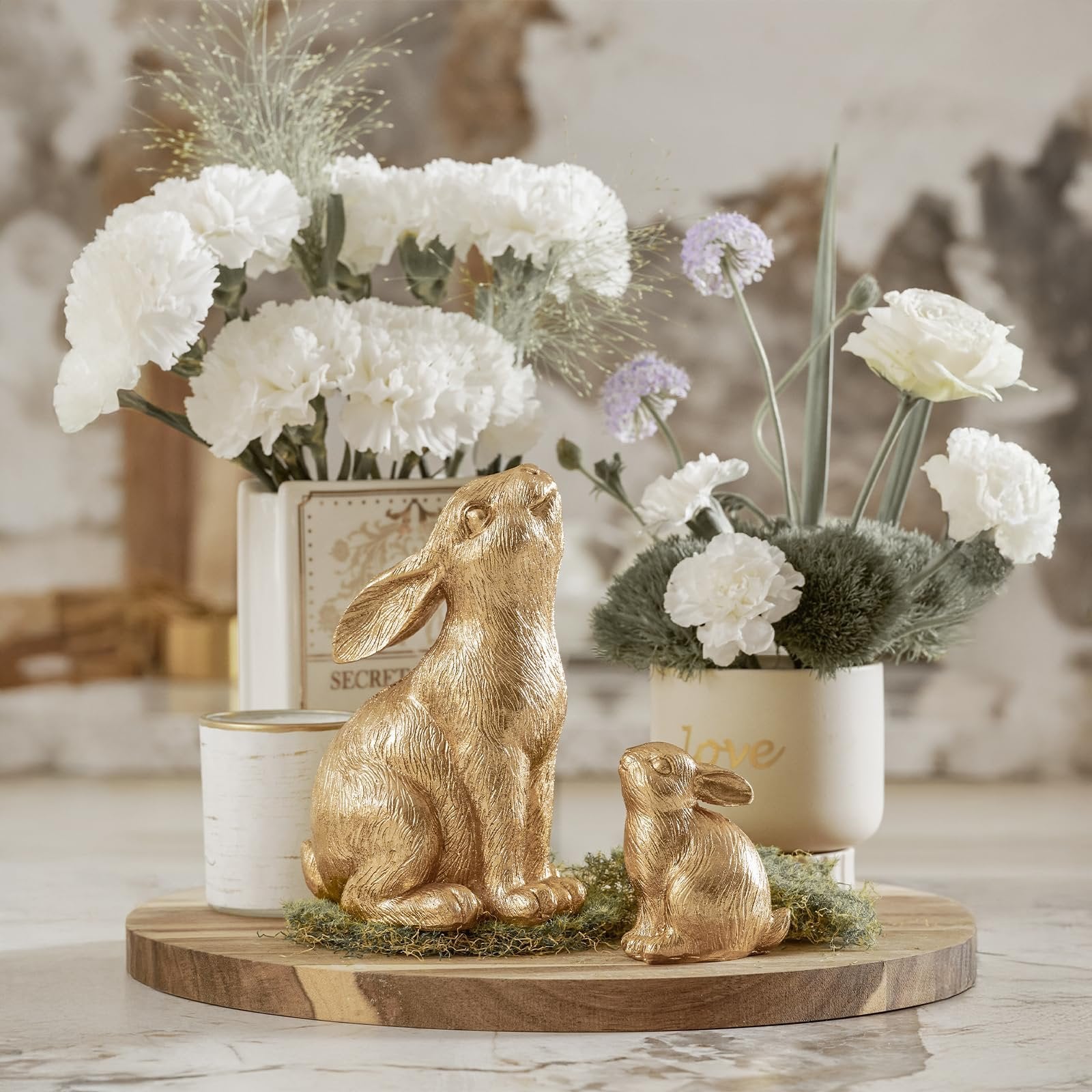 Two gold resin bunny figurines on a wooden board with floral arrangements and decorative vases.