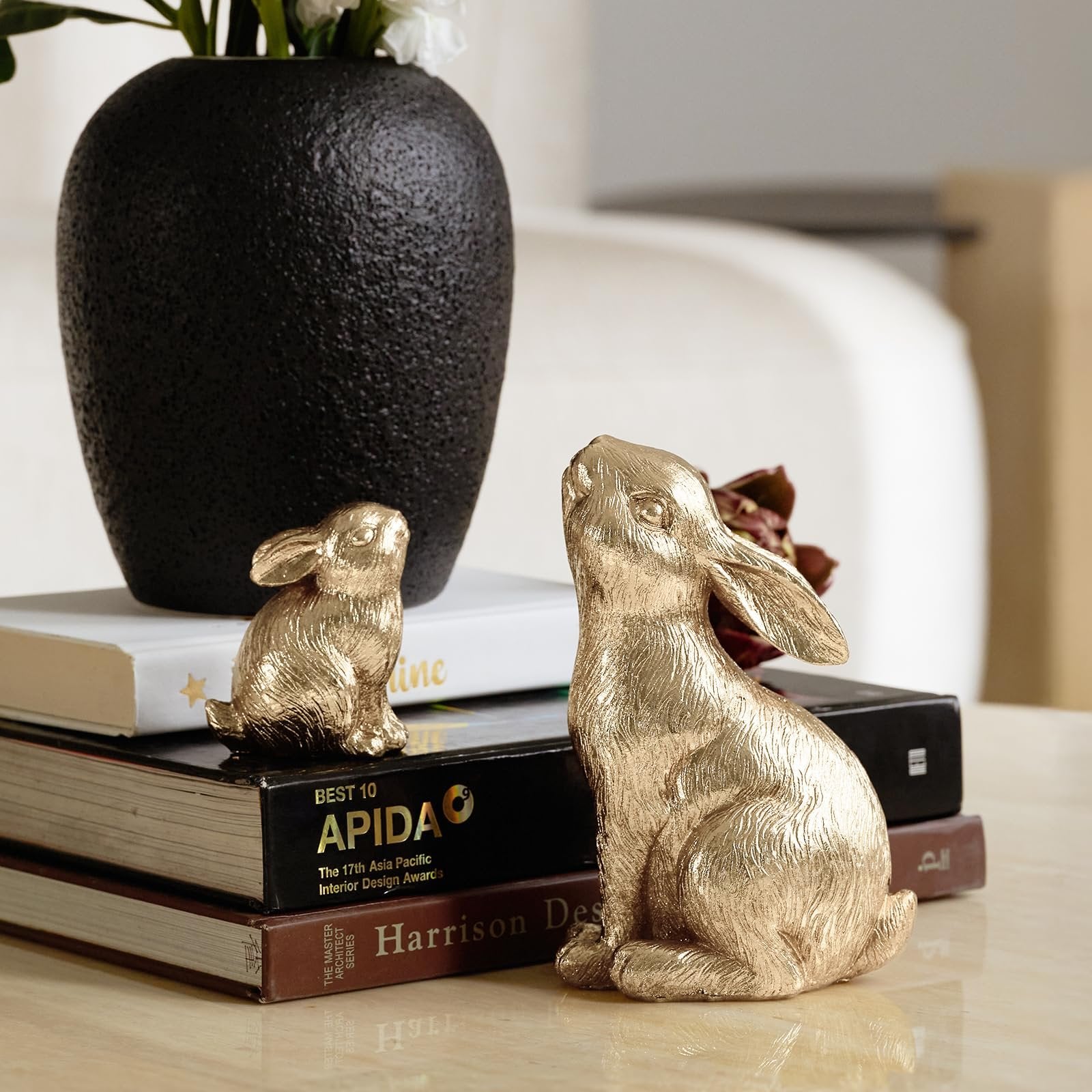 Two gold resin bunny figurines sit on a stack of books next to a black vase with white flowers.
