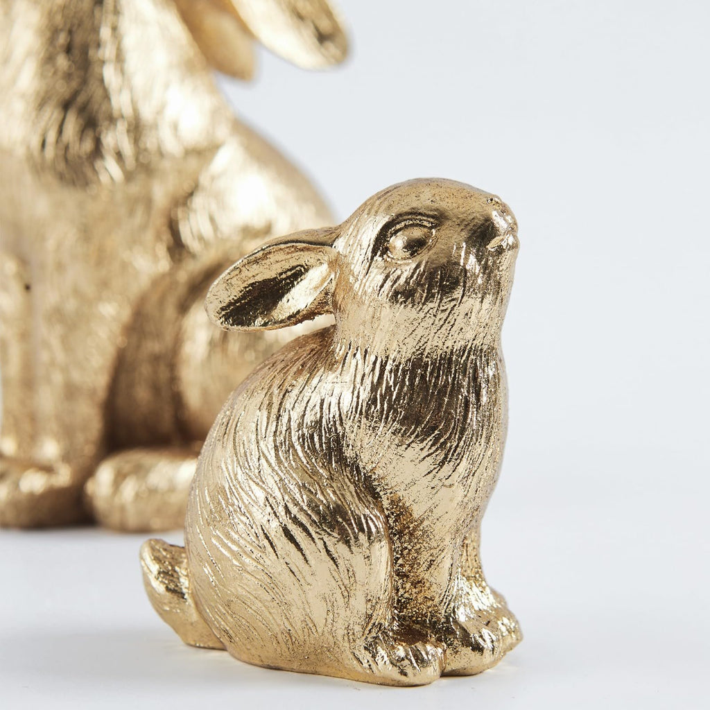 Close-up of a gold resin bunny figurine with textured fur detail, facing right, with a larger blurred bunny in the background on a white surface.