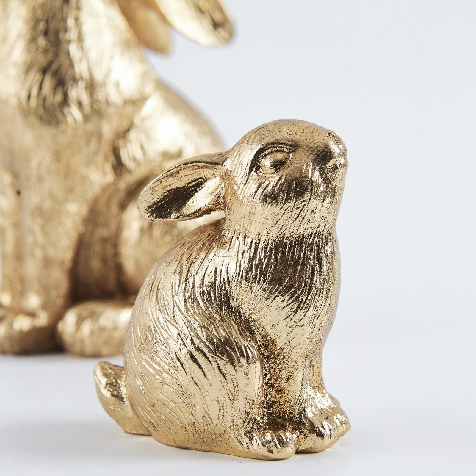 Close-up of a gold resin bunny figurine with textured fur detail, facing right, with a larger blurred bunny in the background on a white surface.