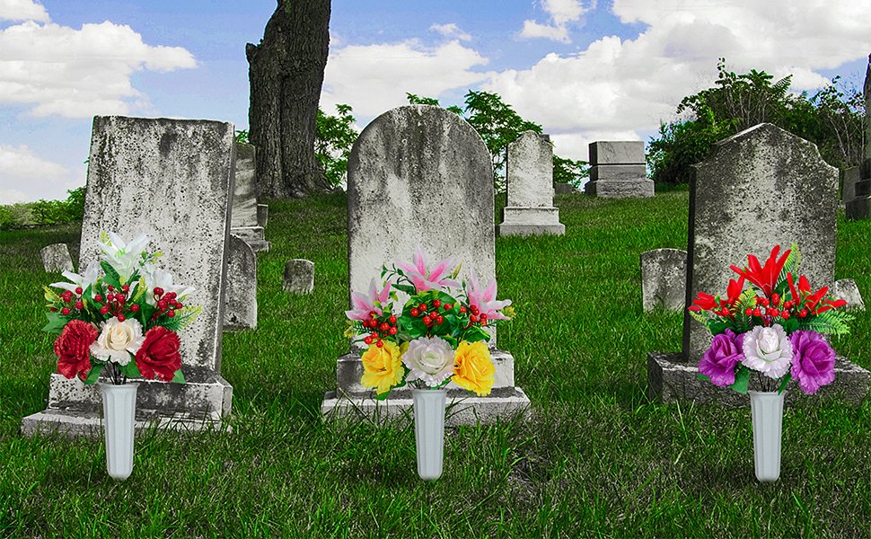 Three artificial flower bouquets in white vases placed in front of gravestones in a cemetery.