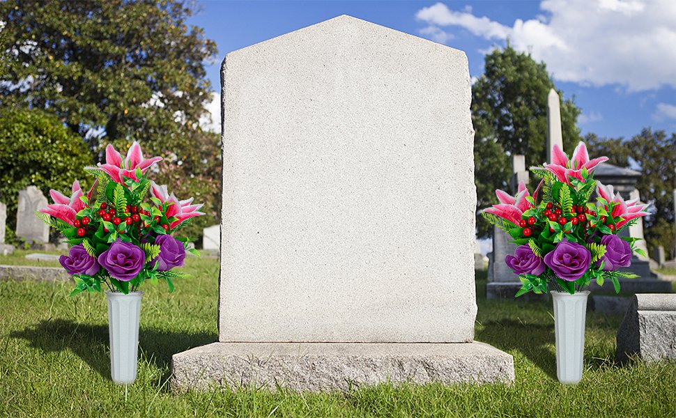 Two artificial flower bouquets with pink lilies and purple roses in white vases placed on either side of a blank tombstone in a cemetery.
