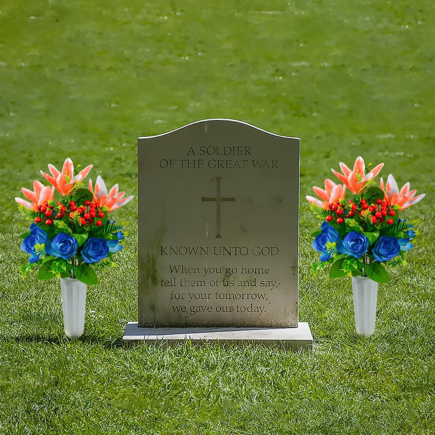 Two artificial flower bouquets with lilies, roses, and berries in white vases placed on either side of a tombstone in a grassy field.