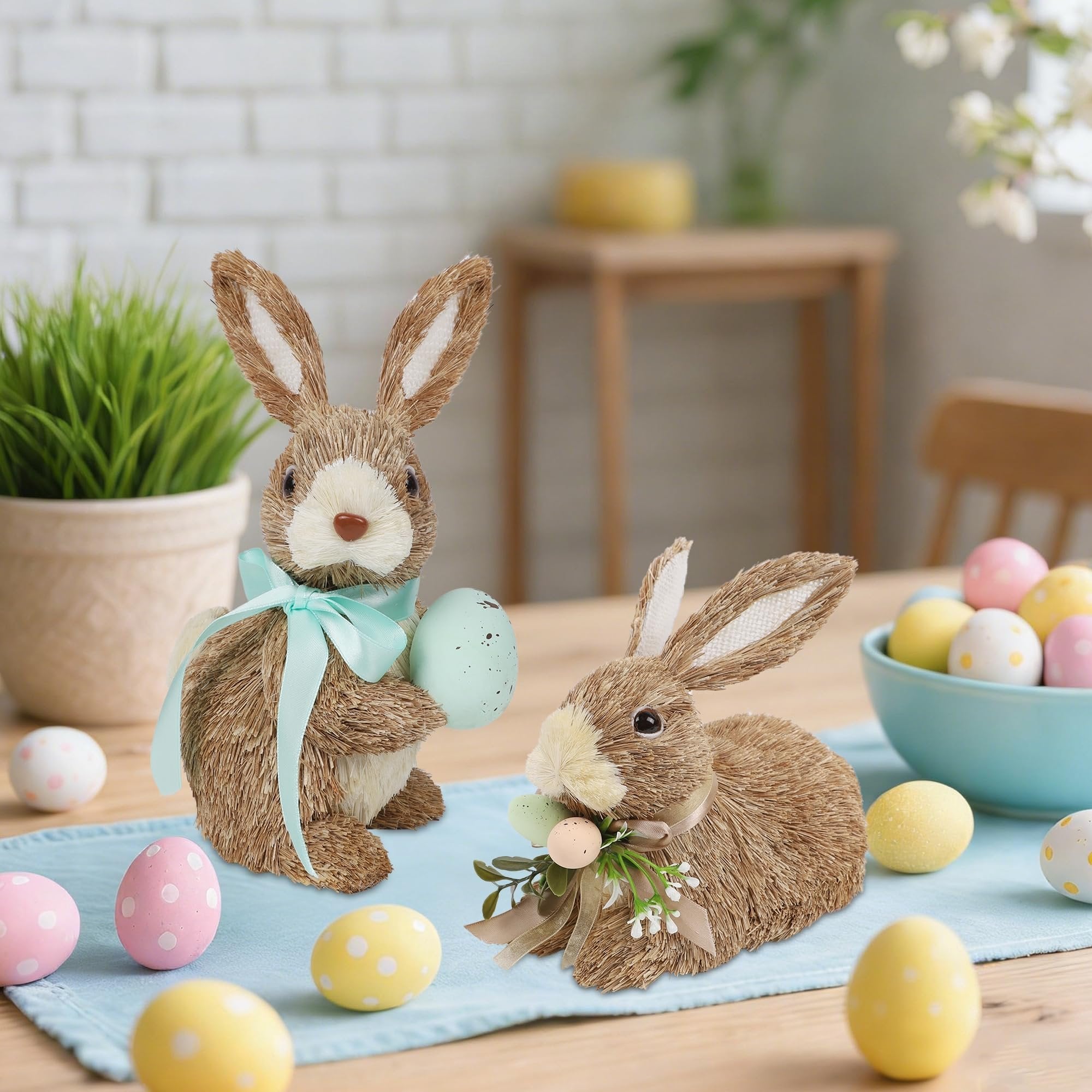 Two sisal bunny figurines with Easter eggs and ribbons on a table, with a bowl of eggs and a potted plant in the background.