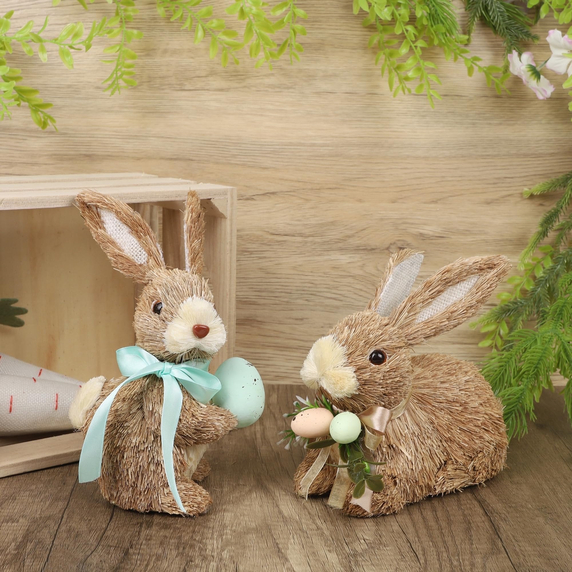 Two sisal bunny figurines with Easter eggs and ribbons on a wooden surface, with greenery and a wooden crate in the background.
