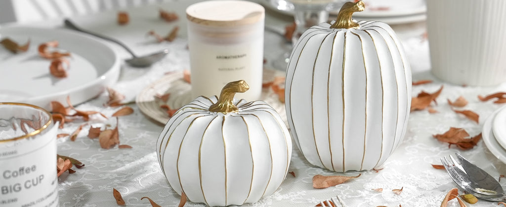 Two white and gold decorative pumpkins on a lace tablecloth with dried leaves and tableware.