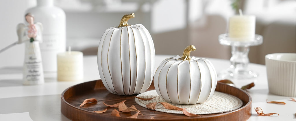 Two white and gold decorative pumpkins on a wooden tray with dried leaves, a candle, and an angel figurine in the background.