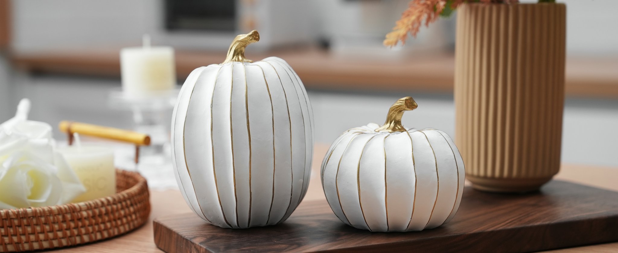 Two white decorative pumpkins with gold stems and accents on a wooden board, with candles and a vase in the background.
