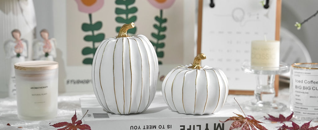 Two white and gold decorative pumpkins on a book, with candles and artwork in the background.