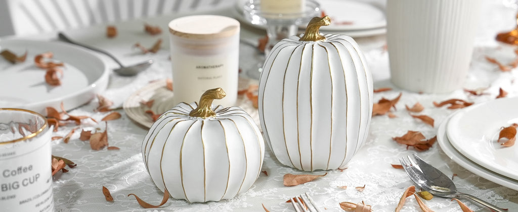 Two white decorative pumpkins with gold accents on a lace tablecloth with dried leaves and tableware.