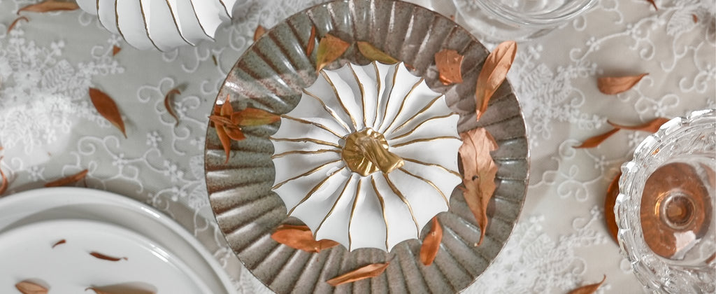 Overhead view of a white and gold decorative pumpkin centerpiece on a textured plate, surrounded by dried leaves.