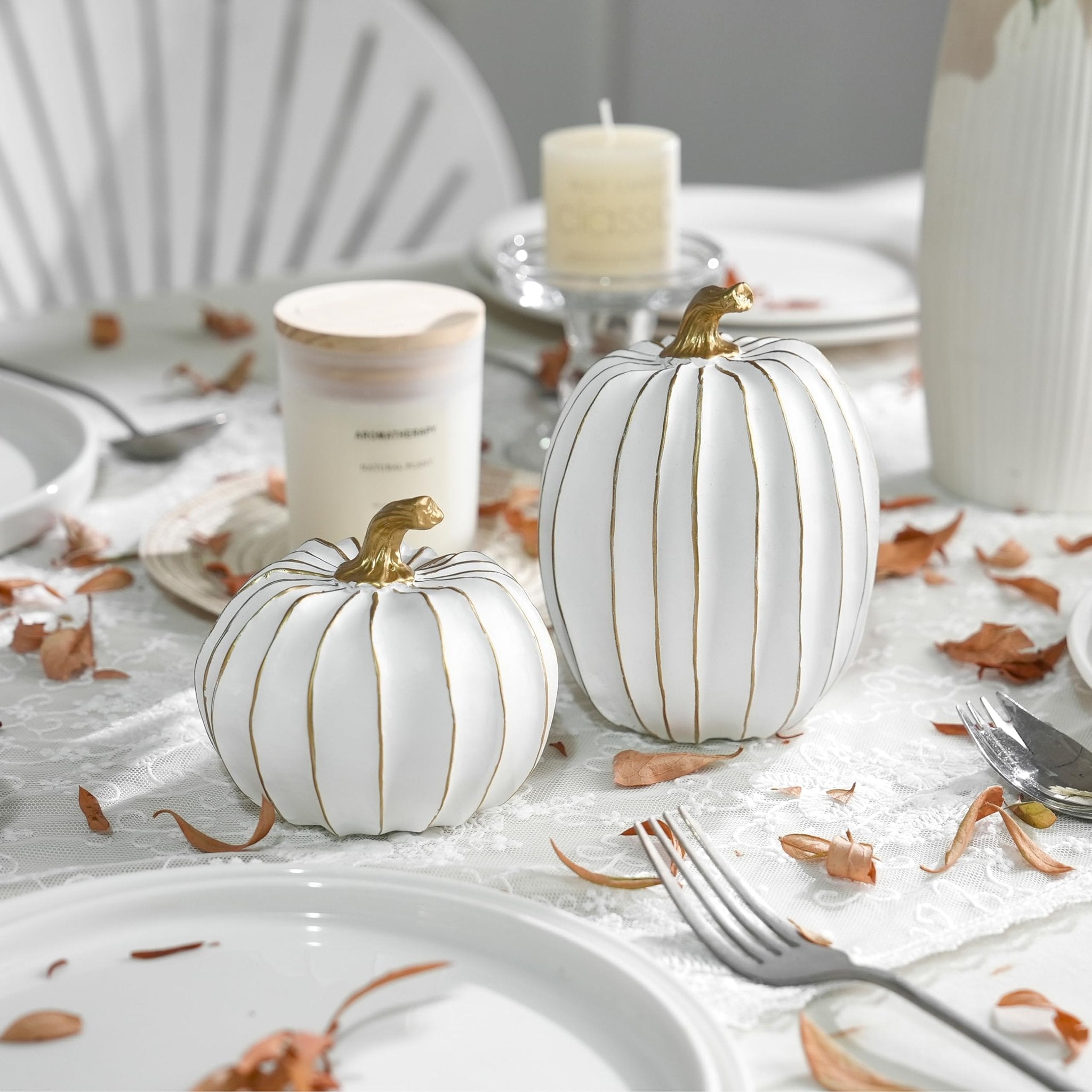 Two white and gold decorative pumpkins on a lace tablecloth with dried leaves and place settings.