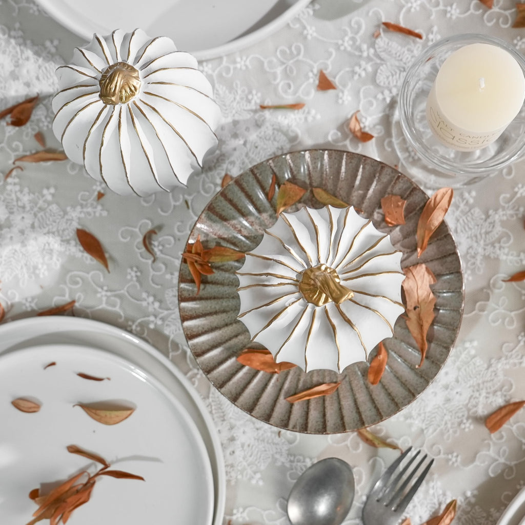 Two decorative white and gold pumpkins on a lace tablecloth with dried leaves and place settings.