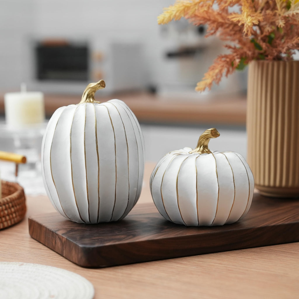 Two white and gold decorative pumpkins on a wooden board with a vase of dried flowers and a candle in the background.