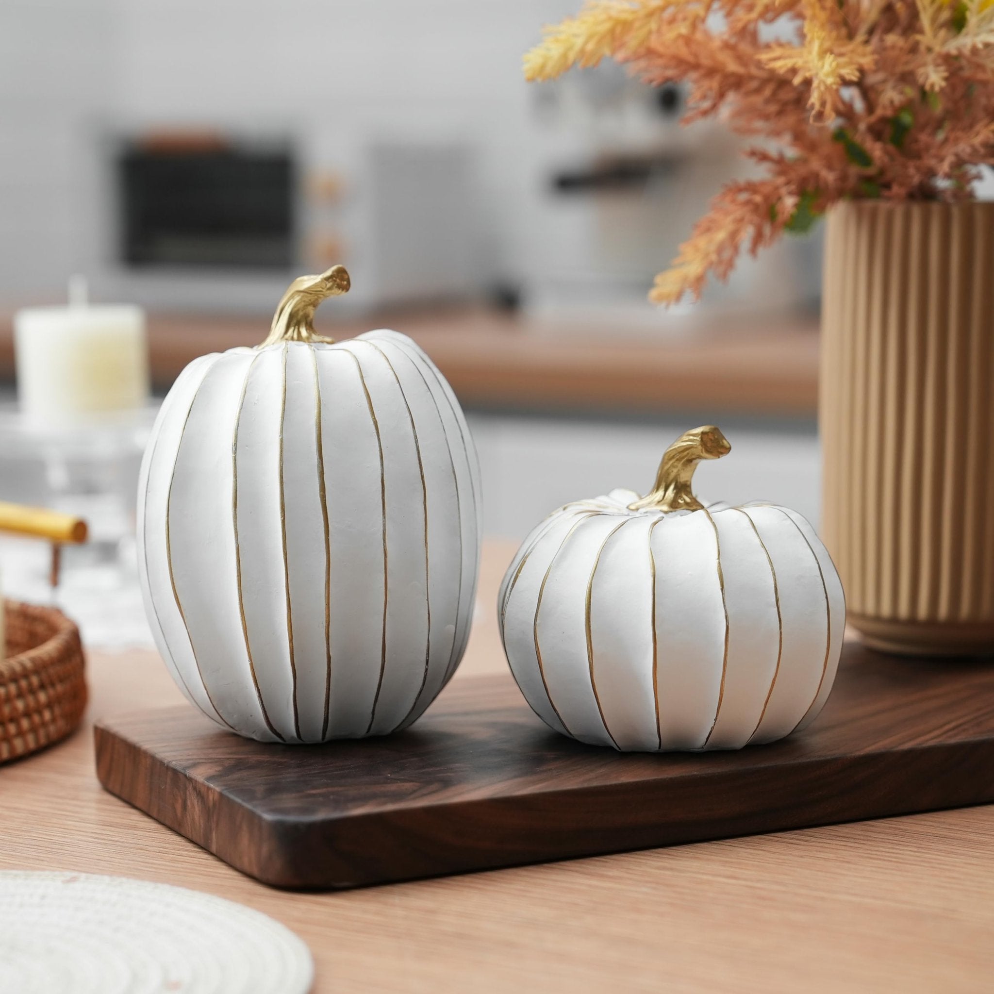 Two white and gold decorative pumpkins on a wooden board with a vase of dried flowers and a candle in the background.