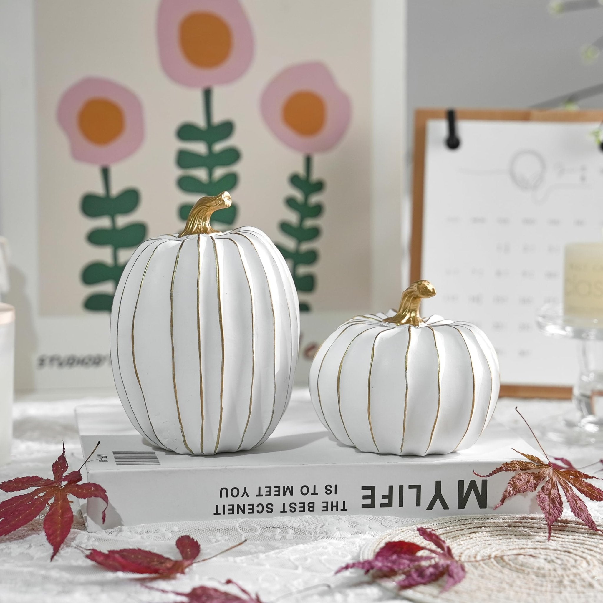 Two white decorative pumpkins with gold stems and gold vertical stripes, arranged on a book with autumn leaves.