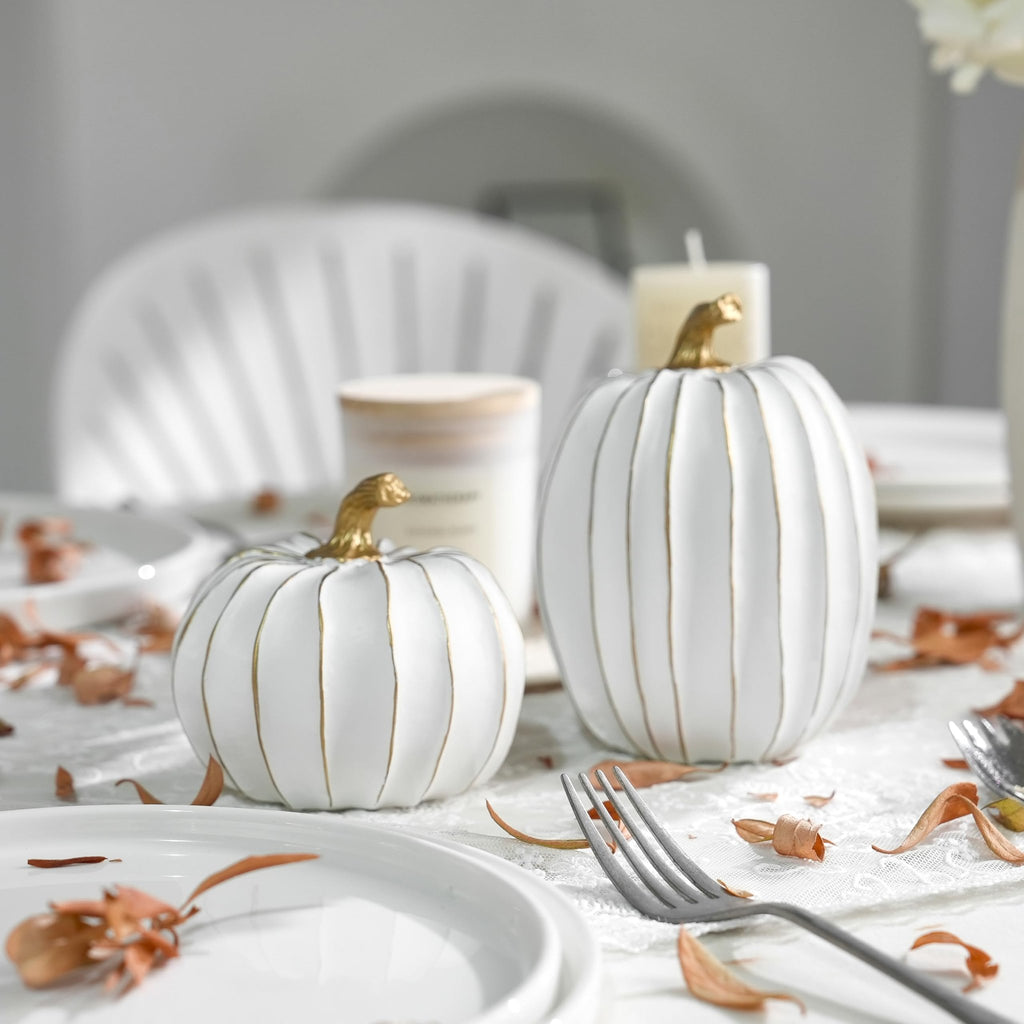 Two white decorative pumpkins with gold stems and accents on a table setting with dried leaves.