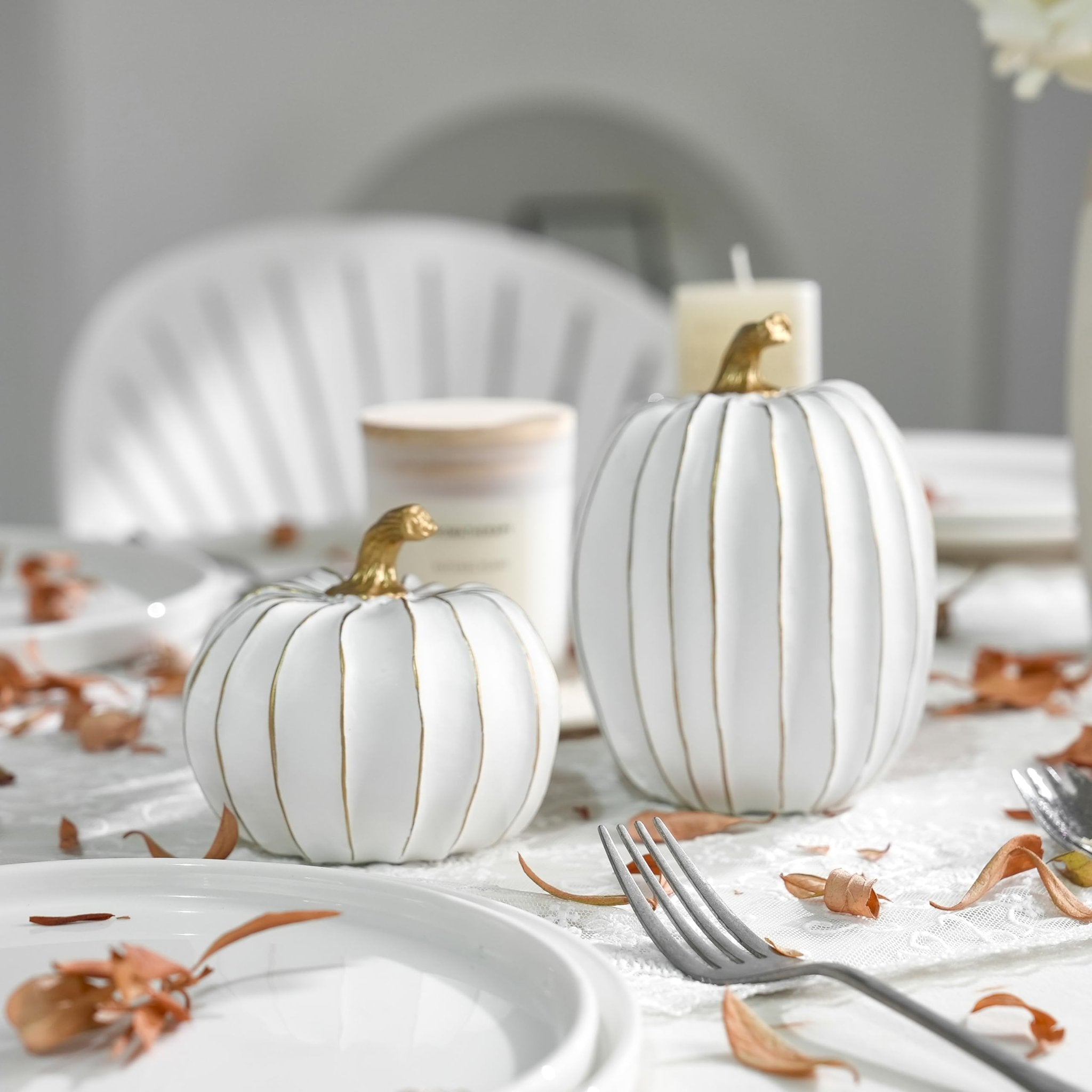 Two white decorative pumpkins with gold stems and accents on a table setting with dried leaves.