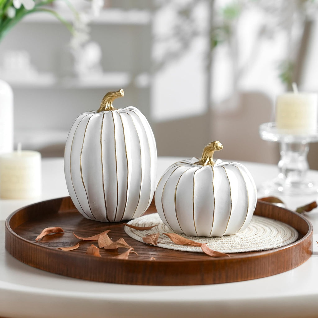 Two white and gold decorative pumpkins on a wooden tray with dried leaves, set on a table with candles and blurred background.
