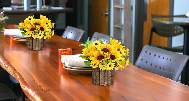 Two potted artificial sunflower arrangements with rustic wood-like vases on a polished wooden table, set for dining.
