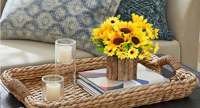 Artificial sunflowers in a rustic pot on a woven tray with books and candles, next to a sofa.