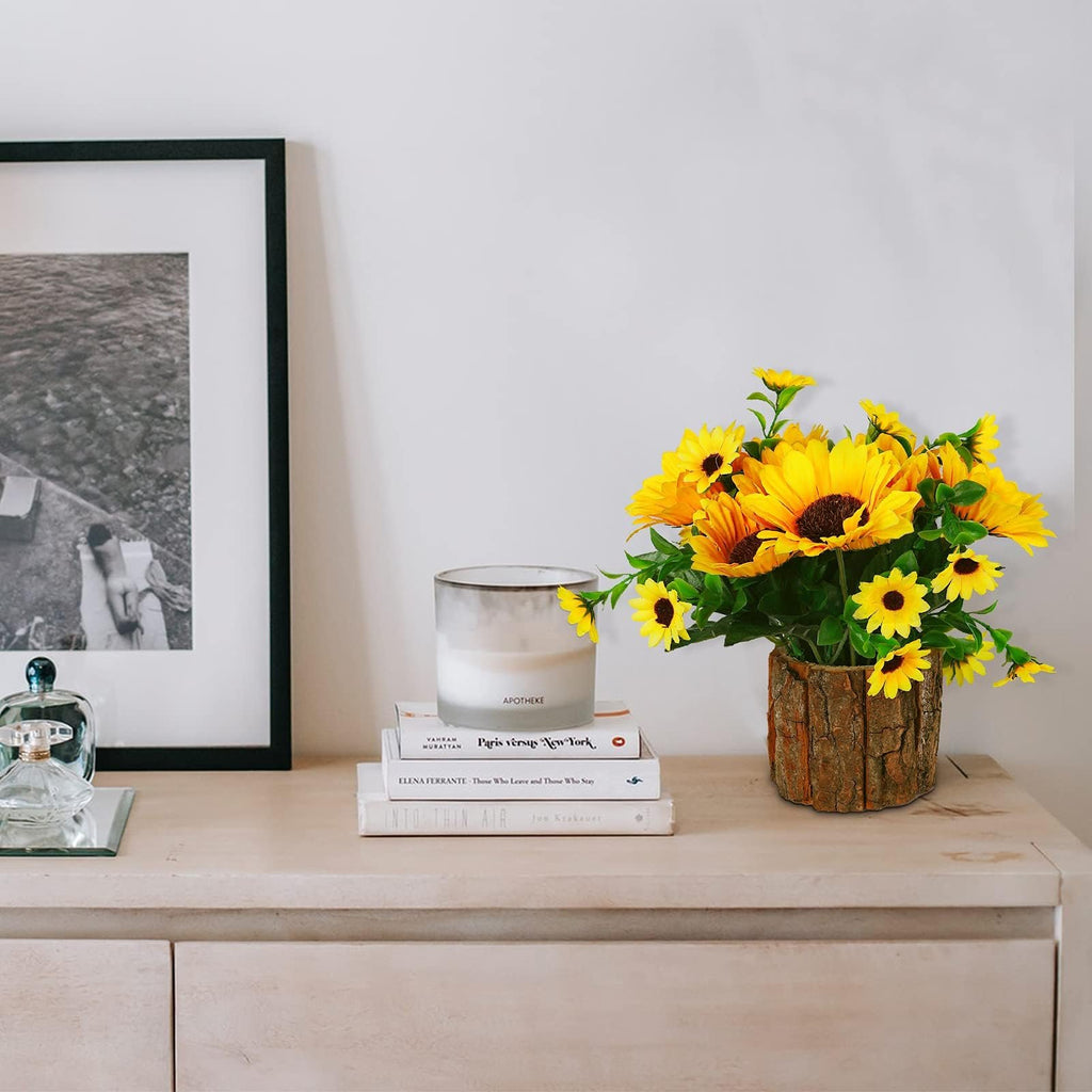 Potted artificial sunflowers with green leaves in a rustic wood-like pot, placed on a shelf next to books and a framed picture.