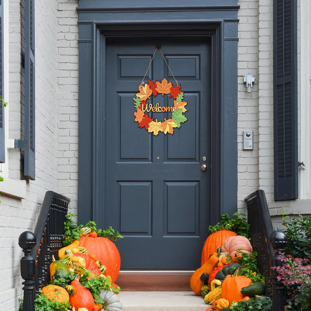 Fall maple leaf wreath with 'welcome' text hanging on a dark blue front door, surrounded by pumpkins and gourds.