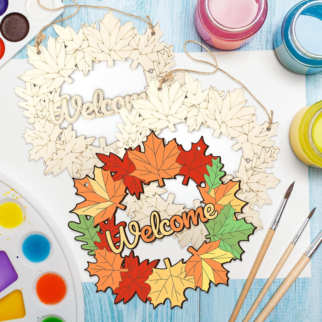 Two unfinished maple leaf wreaths with 'Welcome' cutouts, one with painted autumn leaves, surrounded by paint palettes and brushes.