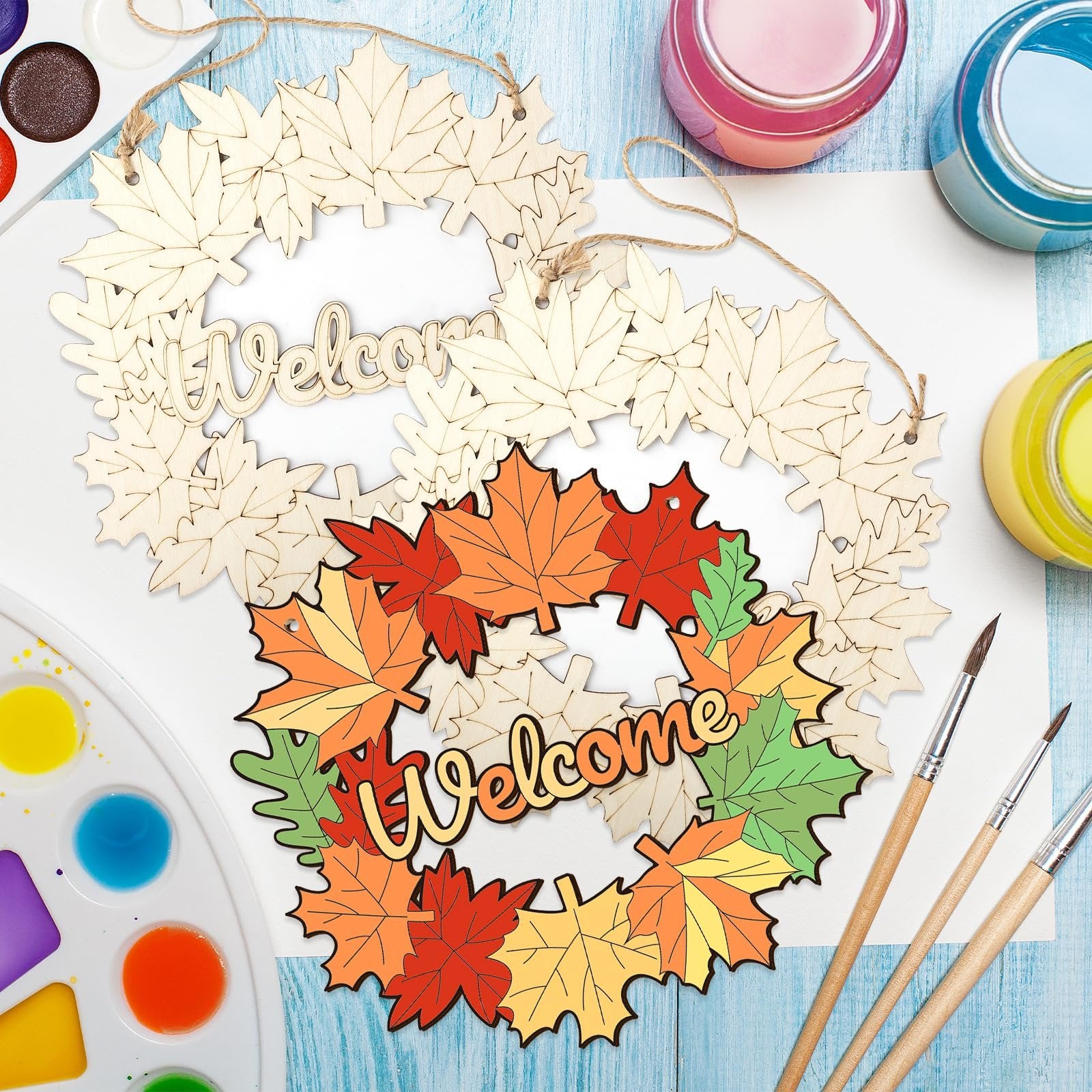 Two unfinished maple leaf wreaths with 'Welcome' cutouts, one with painted autumn leaves, surrounded by paint palettes and brushes.