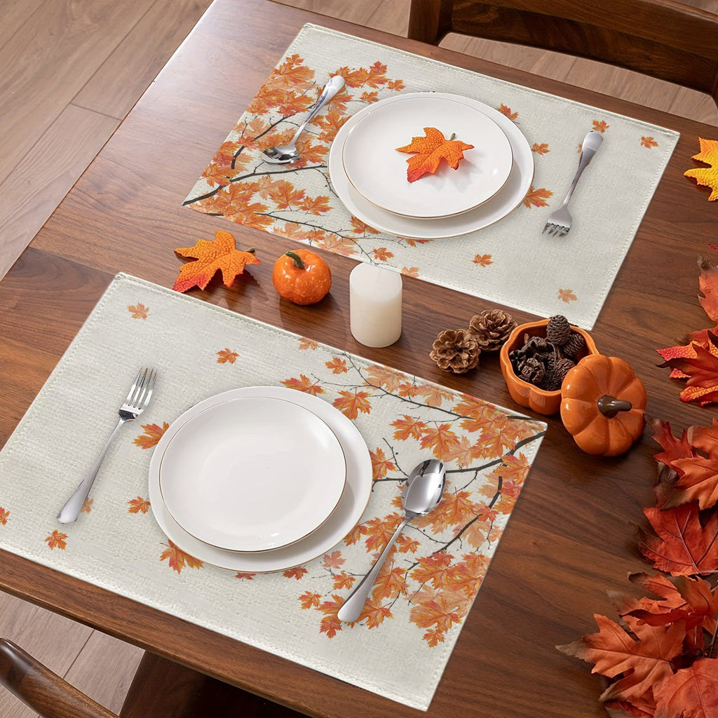 Two fall placemats with autumn maple leaves design set on a wooden table with plates, cutlery, and small pumpkins.
