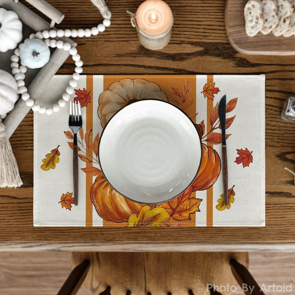 Table setting with a fall pumpkin and leaf placemat, white plate, fork, and knife on a wooden table.