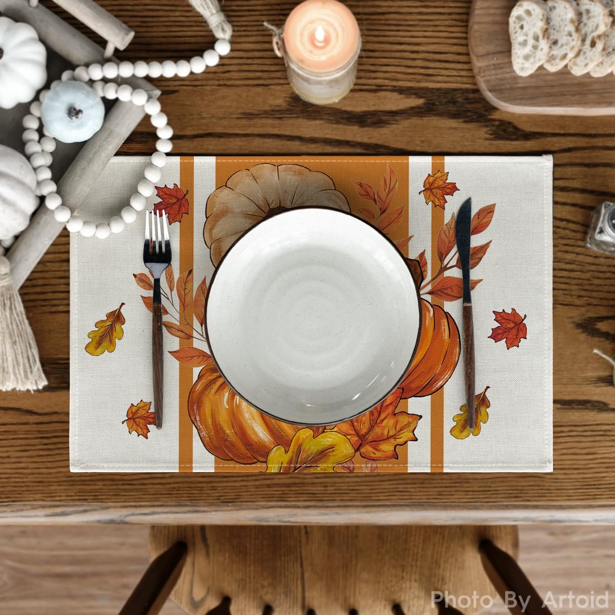 Table setting with a fall pumpkin and leaf placemat, white plate, fork, and knife on a wooden table.