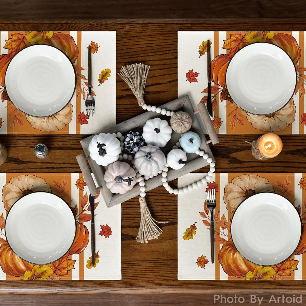 Overhead view of a dining table set with four fall pumpkin leaf placemats, plates, and cutlery, featuring a decorative tray of pumpkins.