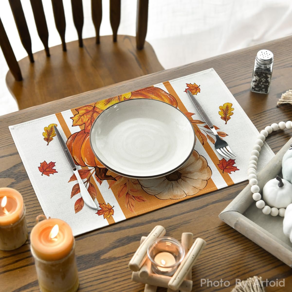 Fall placemat with pumpkins and leaves, set with a plate, fork, and knife on a wooden table with candles.