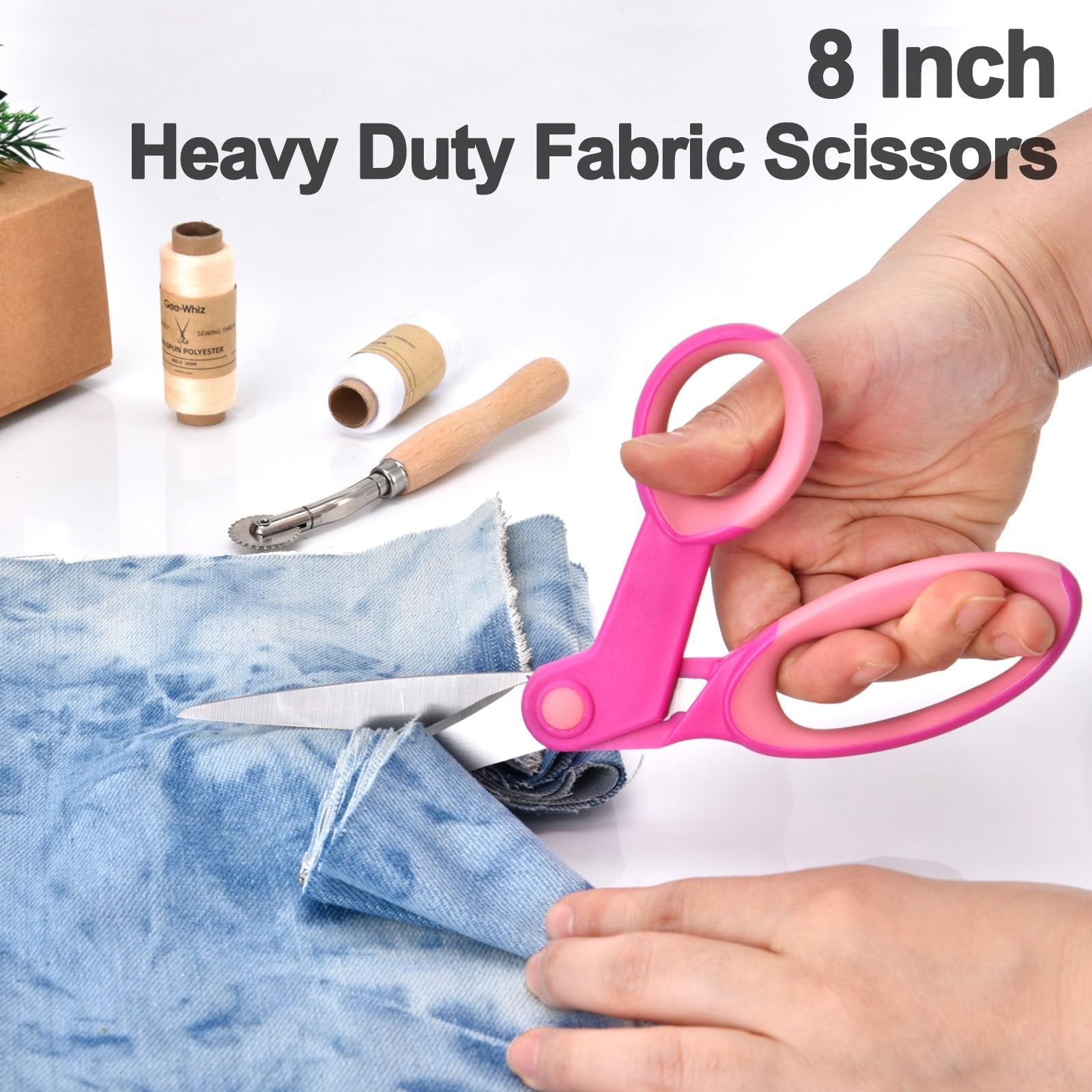 Pink handled 8-inch heavy duty fabric scissors cutting blue denim fabric, with spools of thread and a seam ripper nearby.