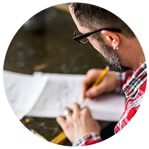 Man with beard and glasses writing in a notebook with a pencil, wearing a red plaid shirt.