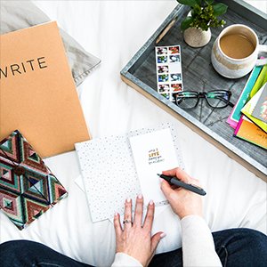 Overhead view of person writing in blank note card with envelopes and journal on bed