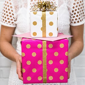 Person in white lace top holding two gift-wrapped boxes, one pink and one white with gold polka dots and ribbon.